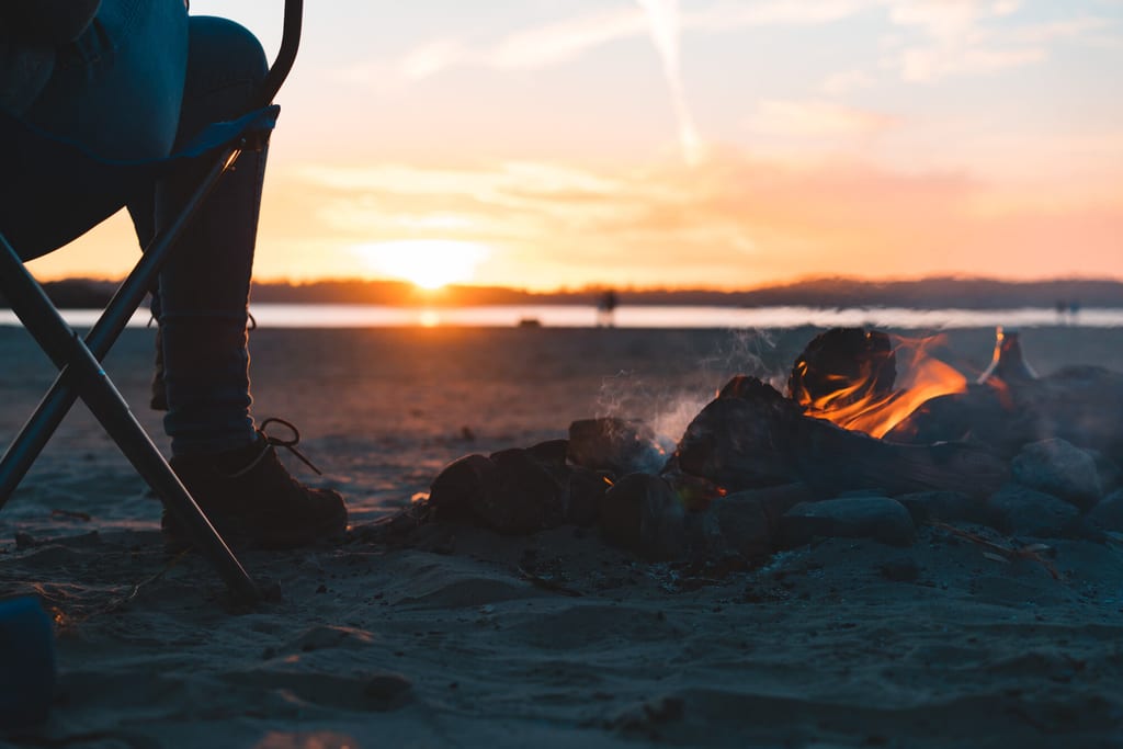 Lagerfeuer am Strand bei Sonnenuntergang, Person sitzt auf Klappstuhl im Vordergrund.