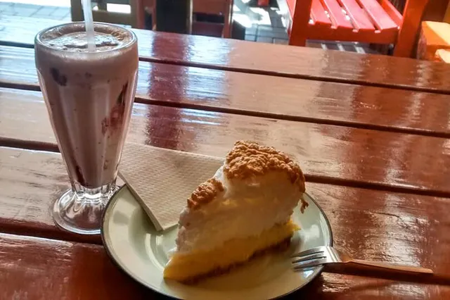 Stück Kuchen mit Baiserhaube und Milchshake auf lackiertem Holztisch in Café-Atmosphäre.