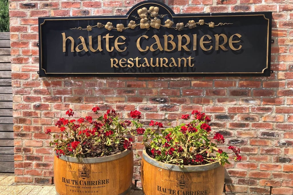 Frontalansicht: schwarzes Restaurantschild an Ziegelwand über zwei hölzernen Fässern mit roten Blumen.