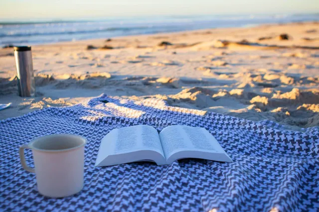 Aufgeschlagenes Buch und Tasse auf einer Decke am Strand bei warmem Seitenlicht.
