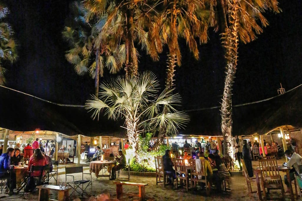 Belebtes Gartenrestaurant unter Palmen bei Nacht mit Gästen an Holztischen