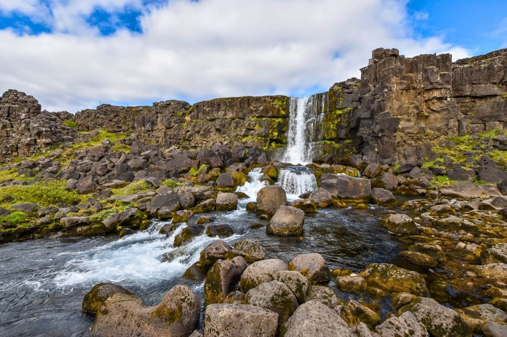 Weitwinkelaufnahme eines Wasserfalls und felsigen Flussufers mit moosbedeckten Basaltsteinen unter bewölktem Himmel.