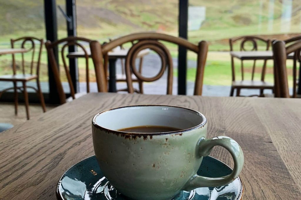 Nahaufnahme einer Kaffeetasse auf Holztisch in Café mit Blick durchs Fenster auf grüne Landschaft.