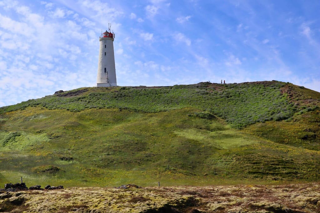 Weißer Leuchtturm auf grasbewachsenem Hügel unter blauem Himmel, zwei Menschen als kleine Silhouetten am Grat.