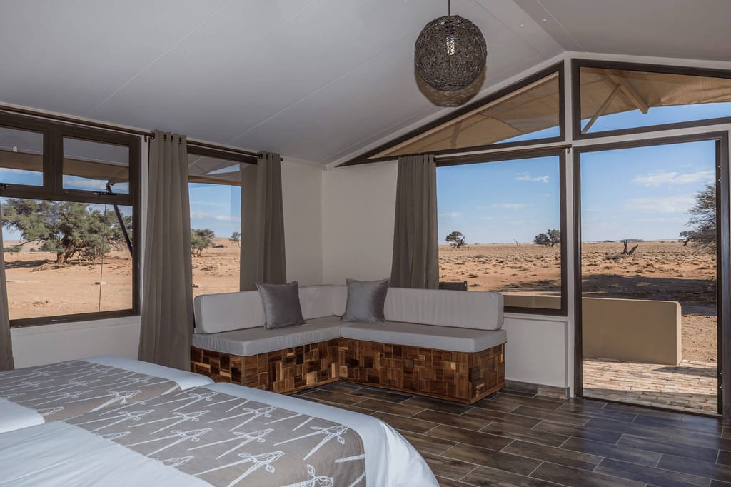 Modernes Schlafzimmer mit großem Fenster und Blick auf trockene Wüstenlandschaft und vereinzelte Bäume.