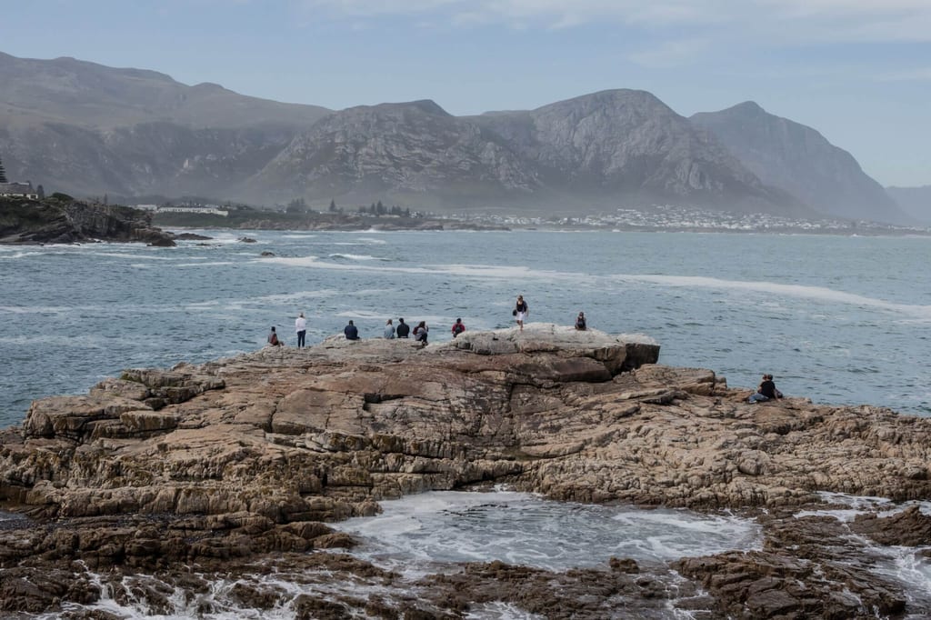 Felsplattform am Meer mit mehreren Personen, Berge im nebligen Hintergrund, bewölkter Tag