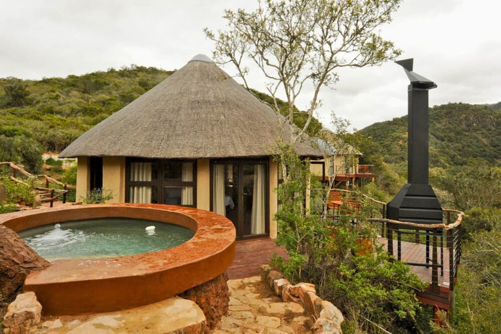 Rundhütte mit Strohdach, Jacuzzi auf Steinplattform und Terrasse mit Kamin in buschiger Hügellandschaft