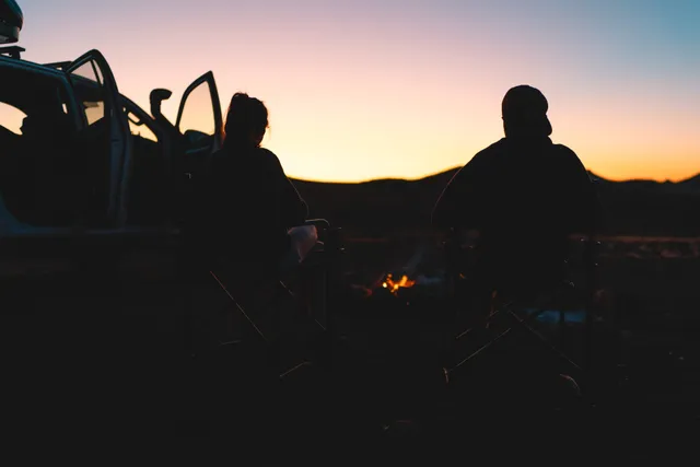 Zwei Personen als Silhouetten sitzen bei Lagerfeuer neben offenem Auto im Sonnenuntergang