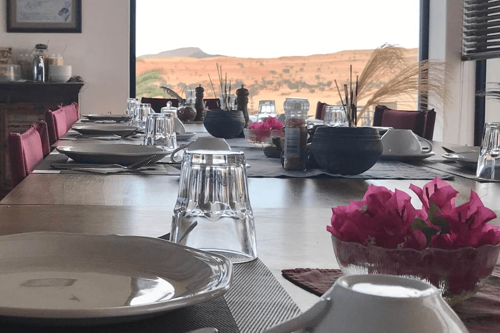 Niedrige Perspektive auf einen lang gedeckten Tisch mit Tellern, Gläsern und rosa Blüten vor sandigen Dünen