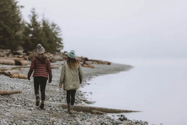 Zwei Personen in Winterkleidung gehen am kiesigen Strand mit Treibholz bei bedecktem Himmel entlang.