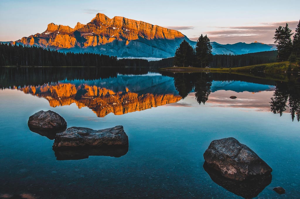 Berg im orangen Licht spiegelt sich in einem ruhigen See, Felsen im Vordergrund.