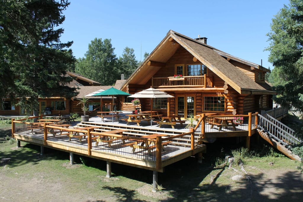Blockhaus mit großer Holzterrasse, Picknicktischen und Sonnenschirmen vor Bäumen und blauem Himmel.