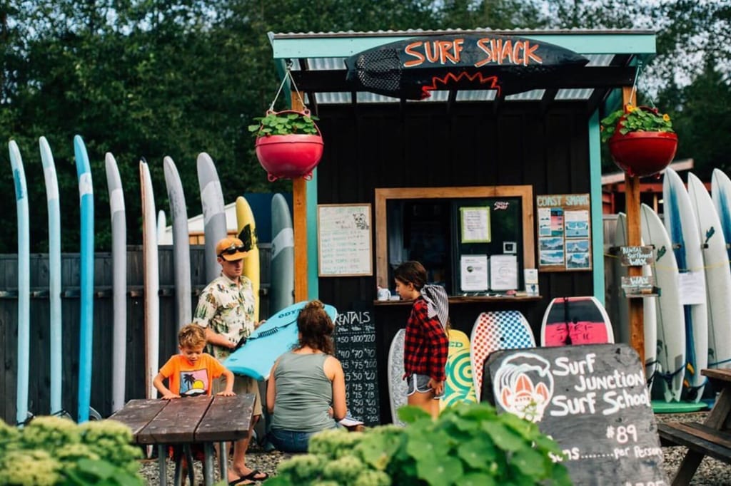Surf-Verleihstand mit aufgereihten Surfboards, mehreren Personen und handbeschildertem Werbeschild in Tageslicht.