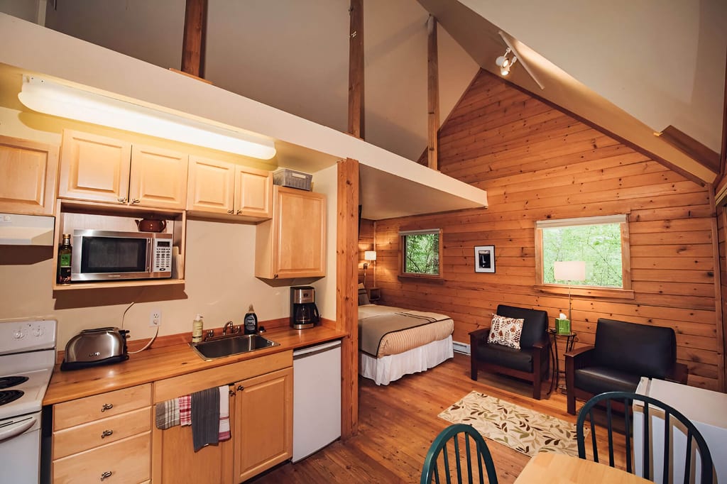 Innenraum einer gemütlichen Holzhütte mit offener Küche, Sitzbereich und Doppelbett, warm beleuchtet.