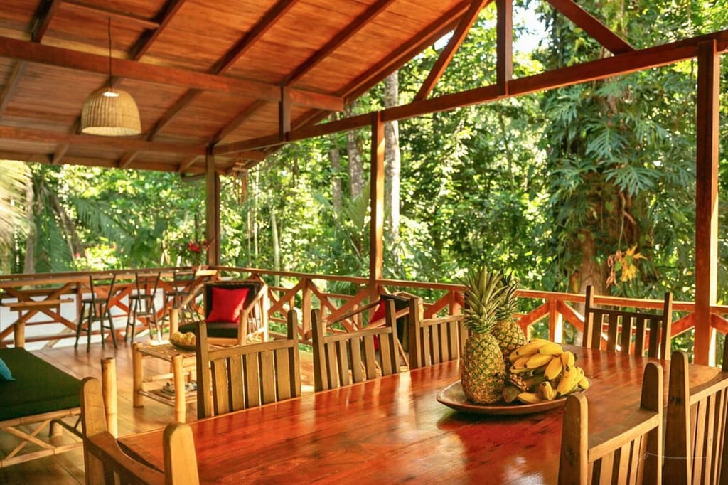 Überdachte Holzveranda mit großem Tisch, Schale mit Ananas und Bananen und Blick in dichten Regenwald
