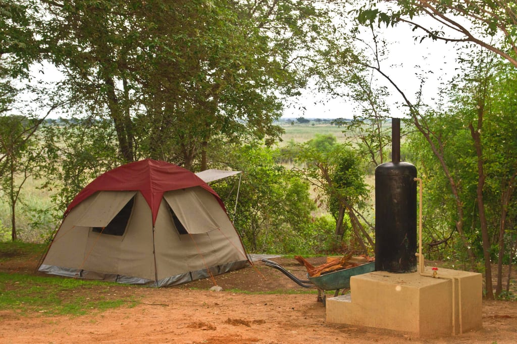 Zeltcamp unter Bäumen mit Schubkarre und schwarzem Zylinder auf Betonsockel, offene Graslandschaft im Hintergrund.