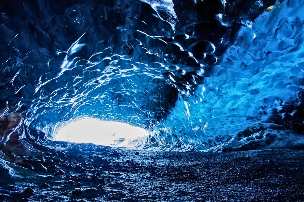 Innenraum einer blau leuchtenden Eishöhle mit heller Öffnung und gerölligem Boden.