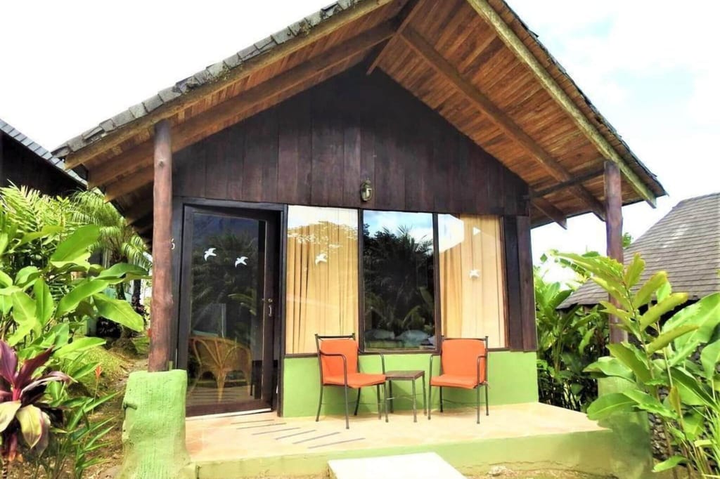Holz-Bungalow mit Veranda, zwei orangen Stühlen und tropischen Pflanzen bei hellem Tageslicht.