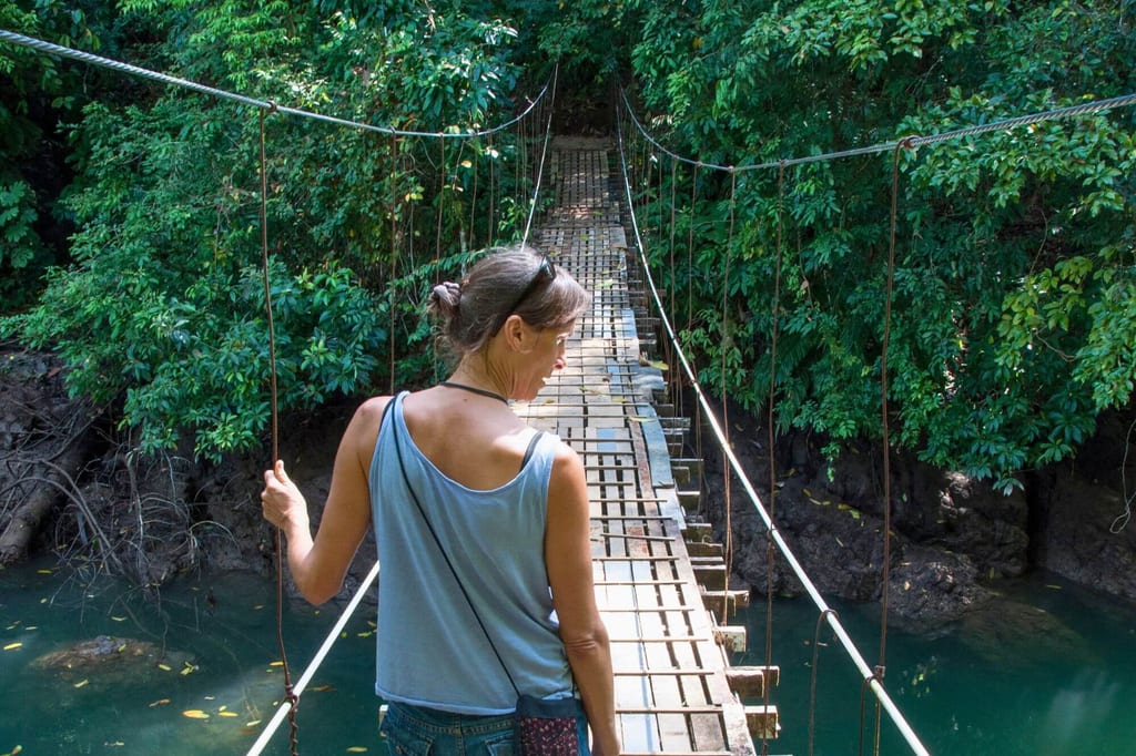 Frau von hinten auf schmaler Hängebrücke über Fluss im dichten grünen Regenwald am Tag.