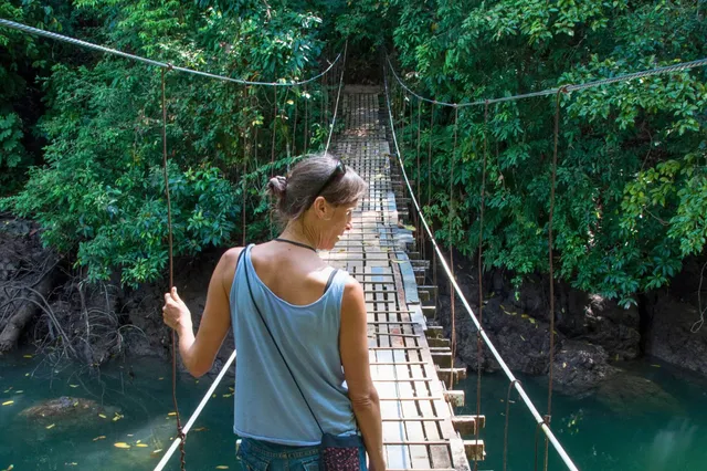 Frau von hinten auf schmaler Hängebrücke über Fluss im dichten grünen Regenwald am Tag.