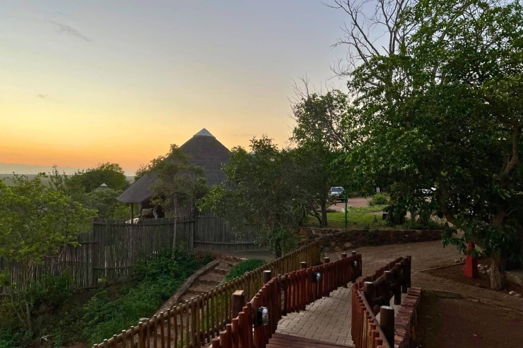 Holzweg führt zu einer Strohdach-Lodge im dichten Grün bei orangem Abendhimmel.