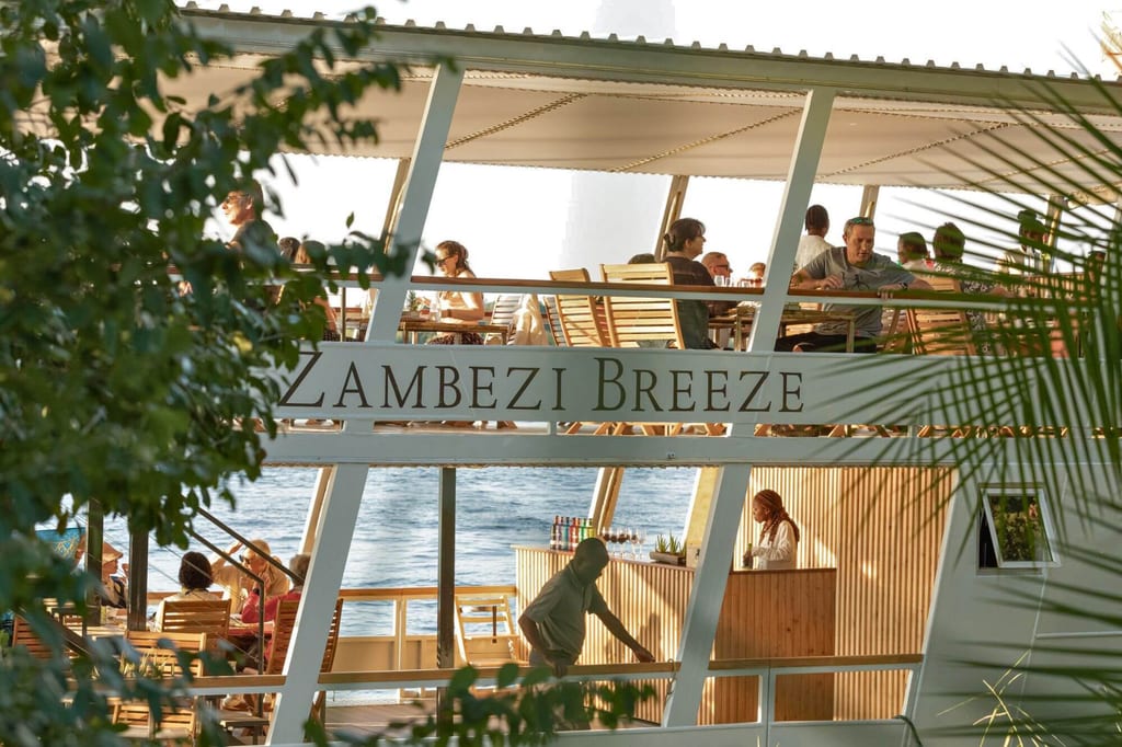 Zweistöckiges Flussboot ‚Zambezi Breeze‘ mit Passagieren auf dem Deck, gesehen durch Ufervegetation.