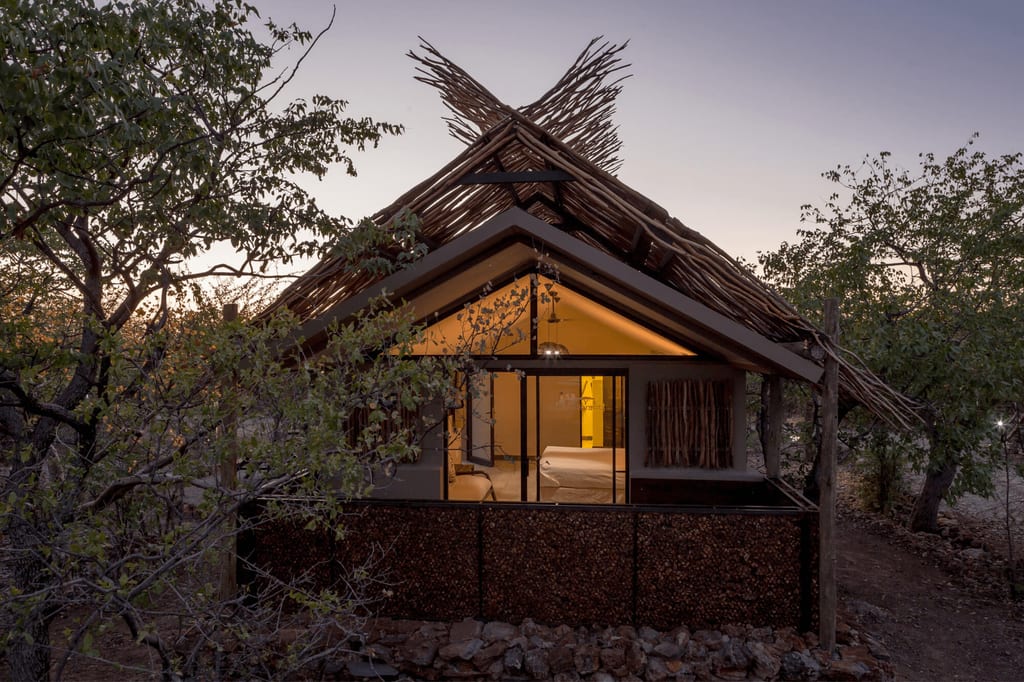 Bush-Lodge mit Strohdach in dichter Buschvegetation bei Dämmerung, warmes Licht innen sichtbar.