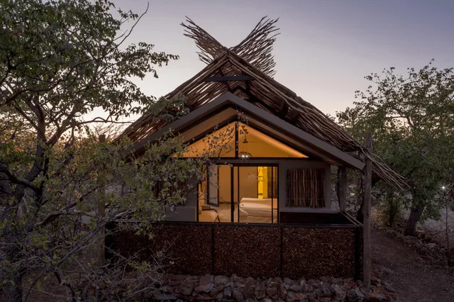 Bush-Lodge mit Strohdach in dichter Buschvegetation bei Dämmerung, warmes Licht innen sichtbar.