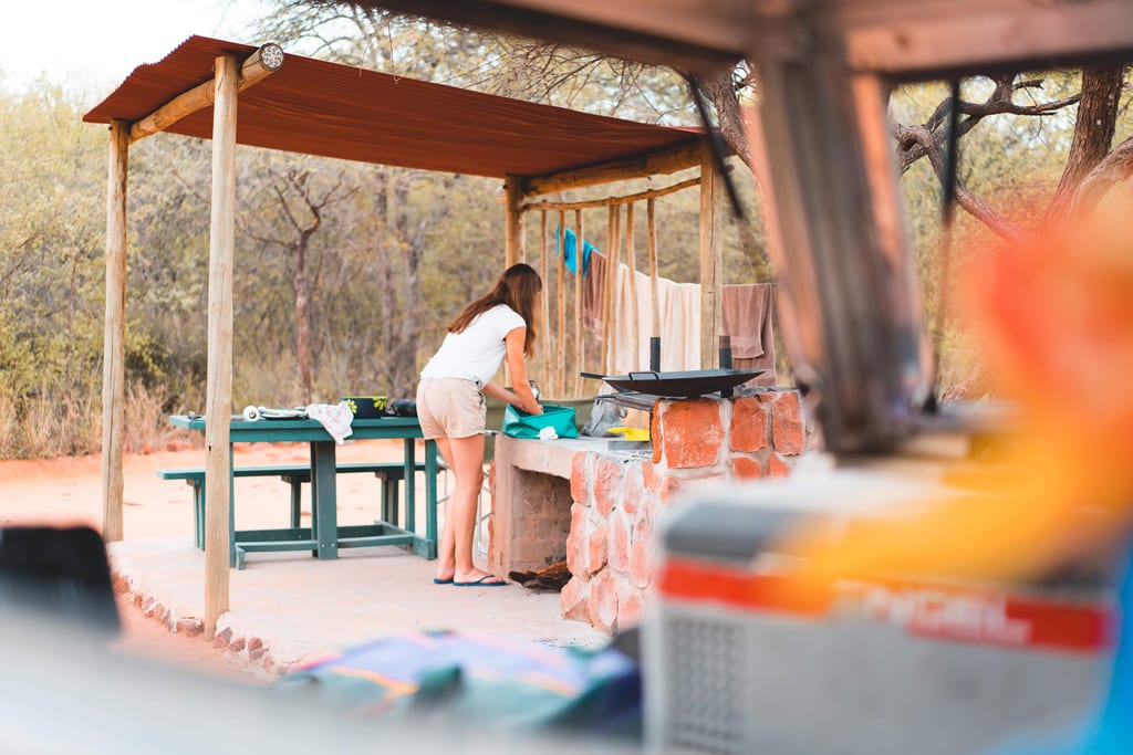 Person beim Abspülen an einer Außenküche auf einem Campingplatz, Handtücher an der Leine, Buschland im Hintergrund.