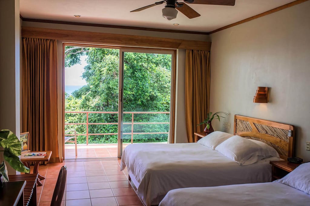 Helles Hotelzimmer mit zwei Betten und Balkon zur grünen Baumkulisse