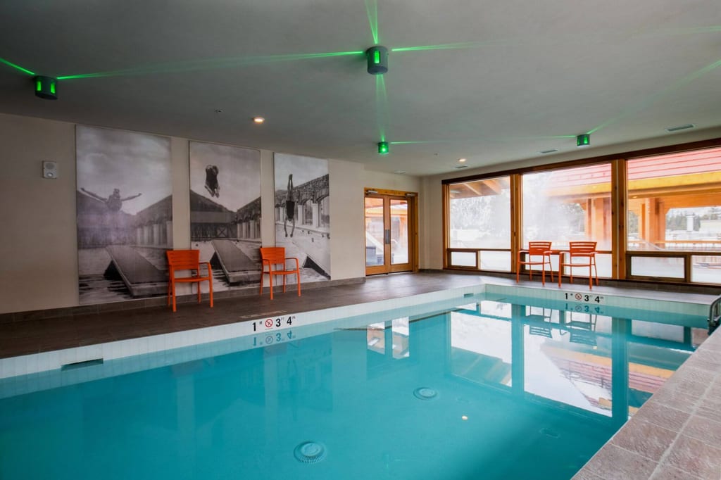 Rechteckiges Hallenbad mit klarer Wasseroberfläche, großen Fenstern und orangen Stühlen.