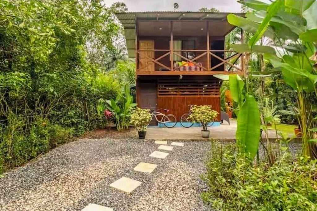 Zweistöckiges Holzhaus mit Balkon, Fahrrädern und Kiesweg in üppigem grünen Garten bei bedecktem Himmel.