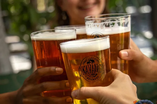 Vier Biergläser mit Schaum werden von Händen zum Anstoßen gehalten, Nahaufnahme in warmem Tageslicht.