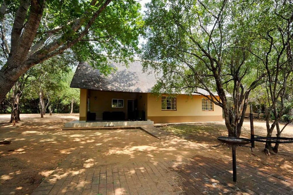Einfache Lodge mit Strohdach und Veranda in schattigem Baumbestand, Sandboden und gepflastertem Weg