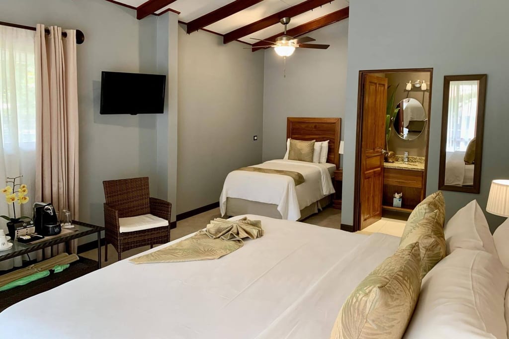 Hell eingerichtetes Hotelzimmer mit Doppelbett, Einzelbett und offenem Bad, Tageslicht von links