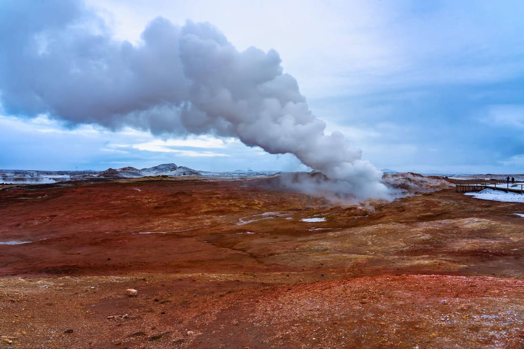 Geothermisches Feld mit rotem Boden und dichter Dampfwolke unter bewölktem Himmel