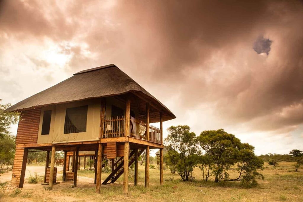 Erhöhte Holzhütte mit Veranda in offener Savannenlandschaft unter dramatischem, bewölktem Himmel.