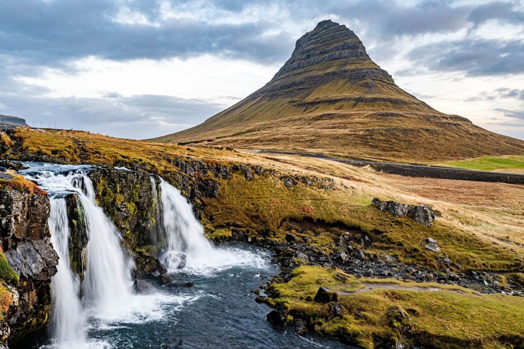 Kegelförmiger Berg und zweigeteilter Wasserfall über herbstlichem Grasland unter bewölktem Himmel