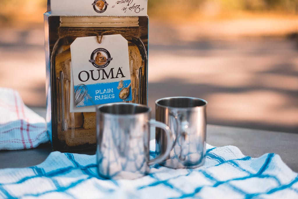Packung OUMA Plain Rusks und zwei Metallbecher auf kariertem Tuch im warmen Abendlicht.