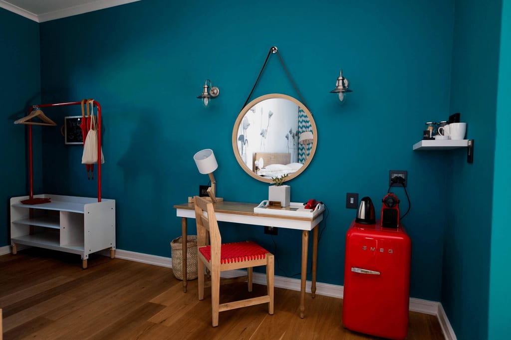 Gemütliches Gästezimmer mit tieftürkiser Wand, Schreibtisch, rundem Spiegel und rotem SMEG-Kühlschrank.