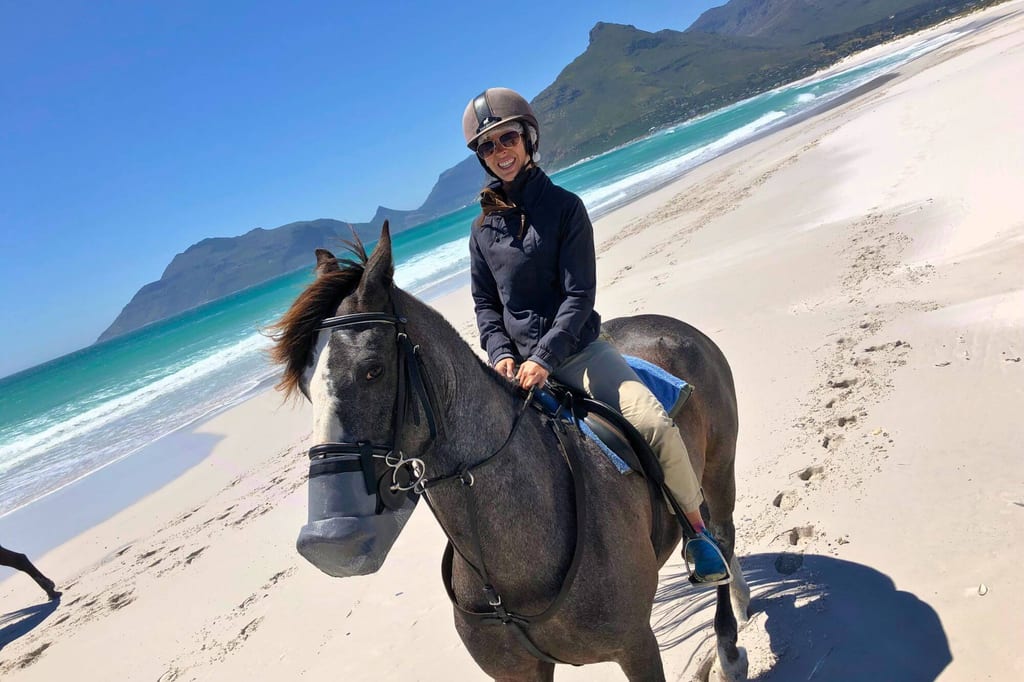 Frau reitet auf dunklem Pferd am sonnigen Sandstrand mit Blick aufs Meer und Berge