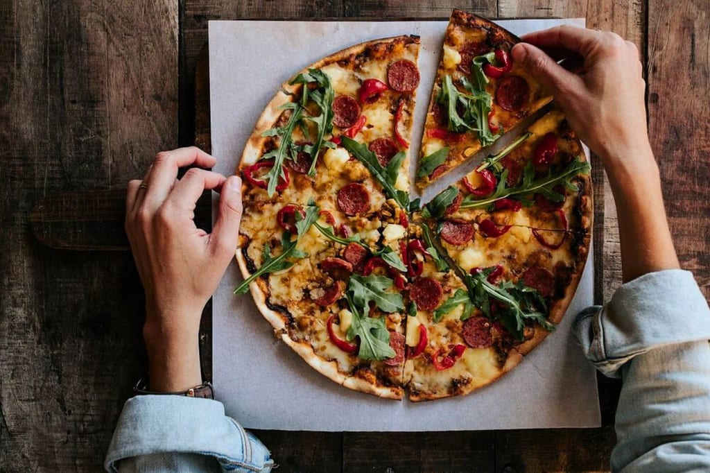 Draufsicht auf eine runde Pizza auf Holztisch, zwei Hände heben ein Stück heraus.