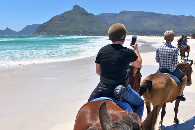 Reiter auf Pferden reiten bei klarem Himmel am Sandstrand mit blauem Meer und Bergen im Hintergrund