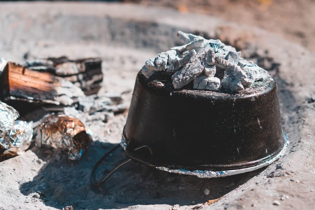 Gusseiserner Topf mit Glut und Alufolien-Paketen in einer Feuerstelle.