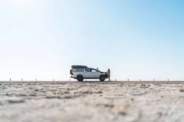 Weißer Geländewagen mit Dachzelt und eine Person auf weiter, flacher Salz- oder Sandfläche unter blauem Himmel.