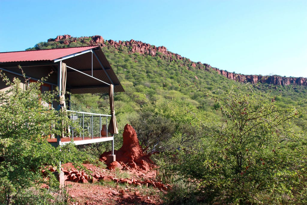 Lodge mit Veranda vor Termitenhügel und grünem Hügelrücken unter klarem blauem Himmel