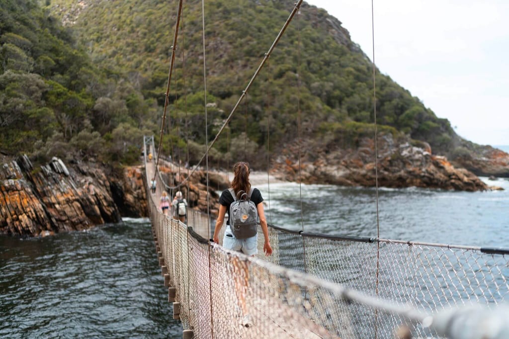 Wanderer mit Rucksack überquert Hängebrücke über Meer vor bewaldeter Felsküste