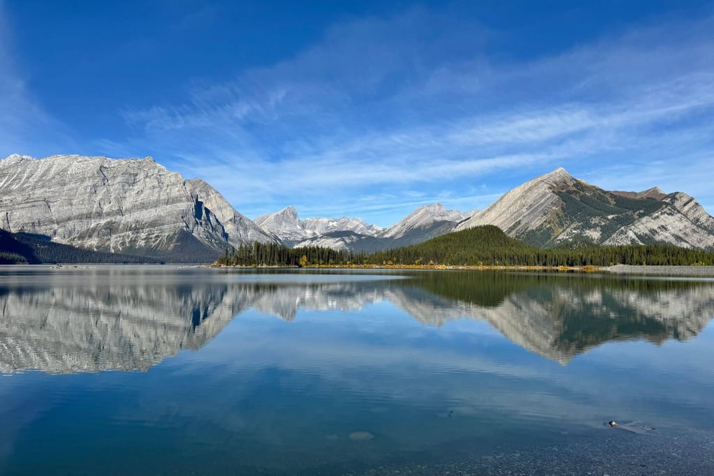 Ruhiger Bergsee mit klarer Spiegelung felsiger Gipfel und bewaldetem Ufer unter blauem Himmel.