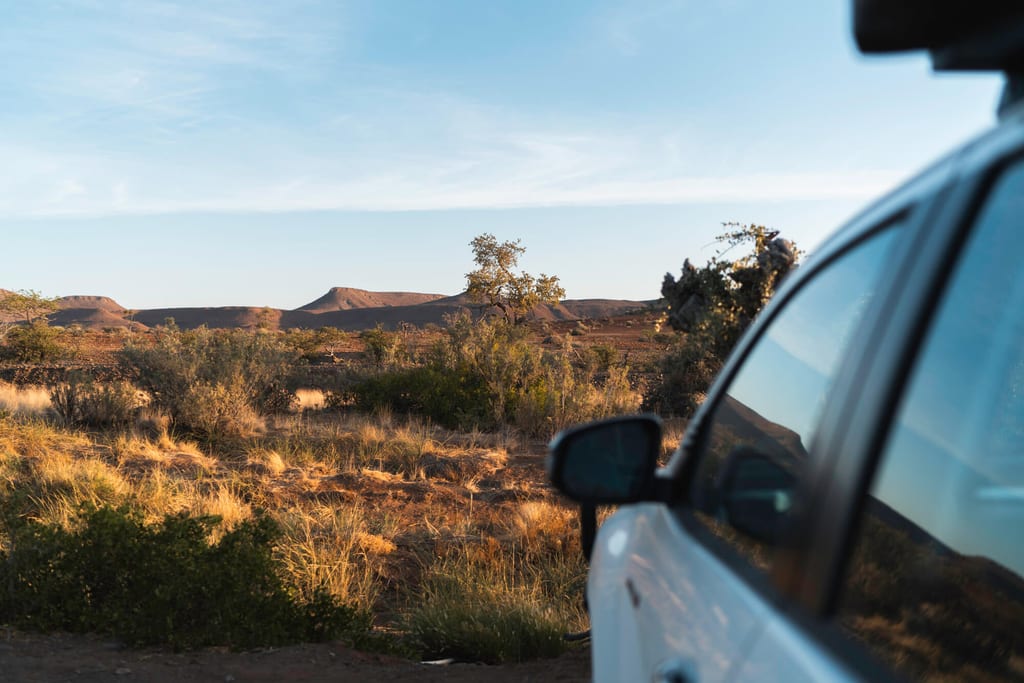 Blick aus dem parkenden Auto über trockene Buschlandschaft zu flachen Tafelbergen im warmen Abendlicht.