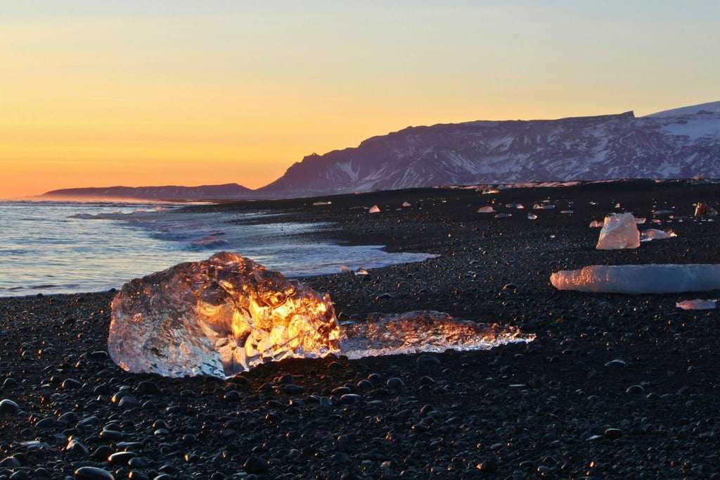 Eisblöcke auf schwarzem Kiesstrand leuchten im warmen Licht des Sonnenuntergangs vor Bergkulisse.